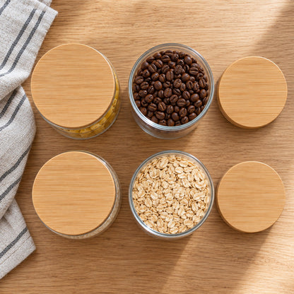 Four glass jars with bamboo lids viewed from above, two open and containing coffee beans and rolled oats
